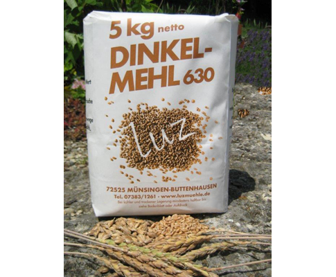 Auf dem Bild ist eine Packung mit der Aufschrift - 5 kg netto Dinkelmehl Type 630 Luz Getreidemühle Münsingen - Buttenhausen - zu sehen, aufgenommen auf einem Stein und im Hintergrund Natur. Vorne liegen geernte Getreidekörner.