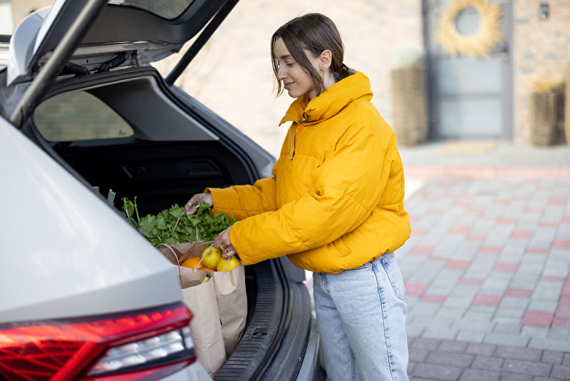 Nah und Gut Bestellen - Abholen von Einkaufsprodukten - Bildinhalt: Junge Frau holt ihre bestellte Ware vom Supermarkt ab. Die Tüten stellt sie in den Kofferraum des Autos.